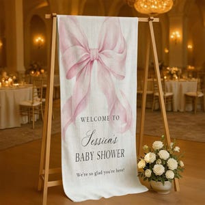 May include: A welcome sign for Jessica's baby shower. The sign features a large, watercolor-style pink bow at the top. The text reads "Welcome to Jessica's Baby Shower. We're so glad you're here!" The sign is displayed on a wooden easel.