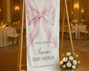 May include: A welcome sign for Jessica's baby shower. The sign features a large, watercolor-style pink bow at the top. The text reads "Welcome to Jessica's Baby Shower. We're so glad you're here!" The sign is displayed on a wooden easel.