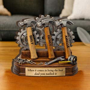 May include: A wooden desk ornament featuring three hammers with personalized names, set against a backdrop of metal gears. The base includes a tape measure, wrench, and a plaque that reads, "When it comes to being the best dad you nailed it."
