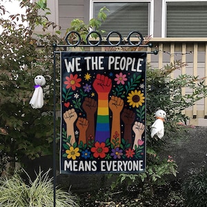 May include: A black garden flag with the text "WE THE PEOPLE MEANS EVERYONE" in white. The flag features raised fists in various skin tones over a rainbow, surrounded by colorful flowers and stars. The flag is displayed on a black metal stand.