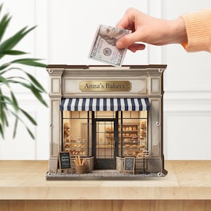 May include: A miniature bakery-themed bank with a blue and white striped awning. The bank has a sign that reads "Anna's Bakery" and a hand is inserting a $100 bill into the slot. The bakery is filled with miniature baked goods.