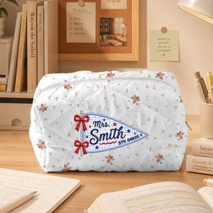 May include: A white quilted pouch featuring a small floral pattern and a central pennant-shaped embroidery. The embroidery displays the text "Mrs. Smith" and "5TH GRADE" in blue, accented by two red bows on the left side and small red stars.