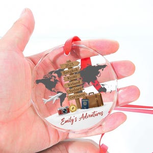 May include: A clear, round ornament with a world map design and travel-themed elements. Features a signpost with city names, a suitcase, passport, camera, and the text "Emily's Adventures". Includes a red ribbon for hanging.
