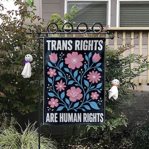May include: A black garden flag with the text "TRANS RIGHTS ARE HUMAN RIGHTS" in white. The flag features pink flowers and blue vines against a black background. The flag is hanging on a black metal stand.