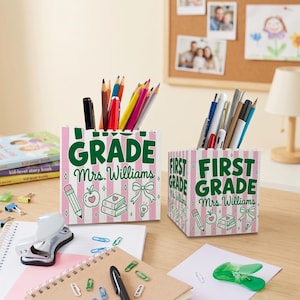 Puede incluir: Dos portalápices cuadrados con un patrón de rayas verticales rosas y blancas y texto verde que dice FIRST GRADE y Mrs. Williams. Los soportes están decorados con ilustraciones de líneas de un lápiz, una manzana, libros y un lazo.