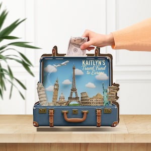 Può includere: Un salvadanaio a forma di valigia blu a tema viaggio. L'interno presenta monumenti famosi come la Torre Eiffel e la Statua della Libertà. Il testo "KAITLYN'S Travel Fund to Europe" è visualizzato. Una mano inserisce una banconota.