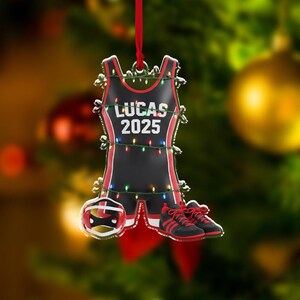 May include: A black and red wrestling singlet Christmas ornament with the name "LUCAS 2025" printed on it. The ornament includes a wrestling helmet and shoes. The ornament is decorated with small lights and a red ribbon for hanging.