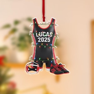 May include: A black and red wrestling singlet ornament with the text "LUCAS 2025". The ornament includes a wrestling helmet and a pair of black and red wrestling shoes. The ornament is decorated with small lights and a red ribbon.