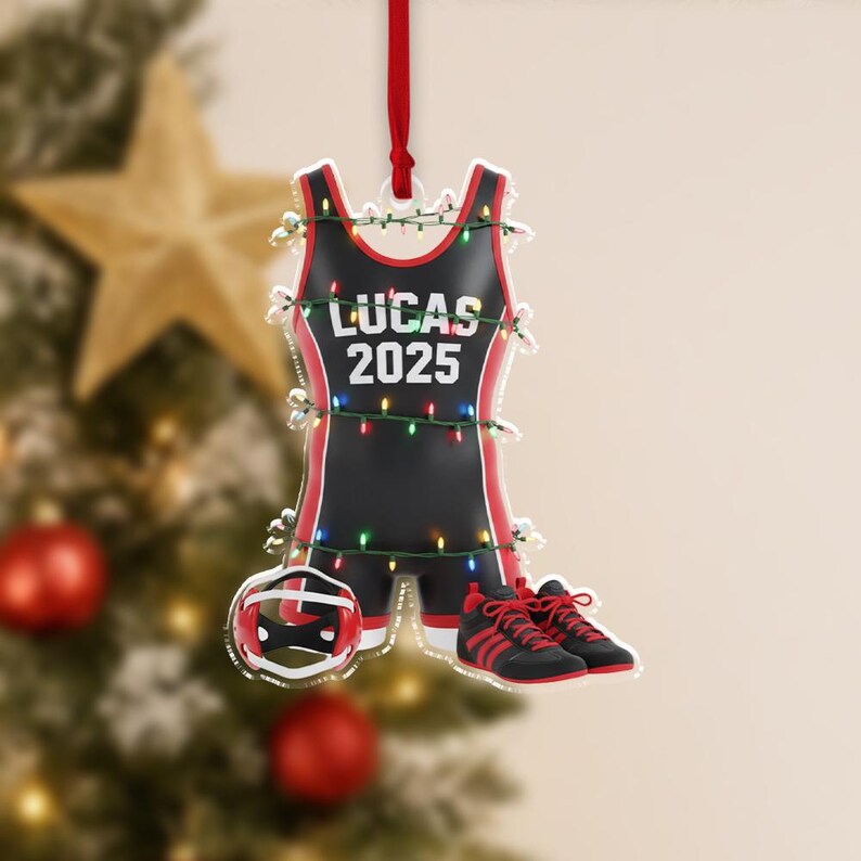 May include: A black and red wrestling singlet Christmas ornament with the text "LUCAS 2025". The ornament includes a wrestling helmet and shoes. The ornament is decorated with small white lights and a red ribbon for hanging.