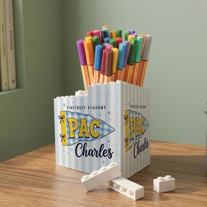 May include: A desk organizer constructed from light blue and white striped building blocks, filled with various colored felt-tip pens. The front features a blue and white checkered pennant graphic with the text Pinecrest Academy, PAC, and Charles.