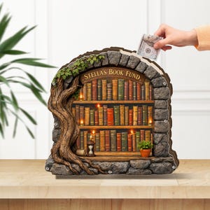 May include: A decorative bookshelf-shaped money bank with the text "STELLA'S BOOK FUND." The bank features a tree, stone accents, and a shelf filled with books. A hand is inserting a dollar bill into the slot.