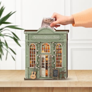 May include: A mint green wooden bank shaped like a library, with the text "Sophia's Library Fund". The bank has detailed windows, a small guitar, and a chalkboard sign. A hand is inserting a £78.90 bill into the slot.