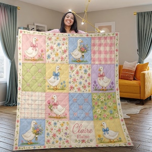 Könnte beinhalten: Eine Patchwork-Decke mit einem sich wiederholenden Muster aus Gänsen, Schleifen und Blumenarrangements in Pastellfarben. Die Decke hat einen floralen Rand und den Namen "Claire Elaine" auf einem Quadrat gestickt.
