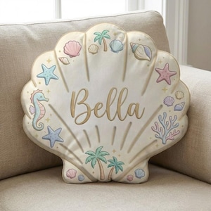 Puede incluir: Un cojín con forma de concha de color crema con el nombre "Bella" en letras doradas. Está adornado con ilustraciones en tonos pastel de conchas marinas, estrellas de mar, caballitos de mar y palmeras. El cojín está sobre un sofá beige.