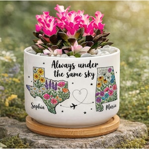 May include: A white ceramic planter with a floral design and the text "Always under the same sky." The planter features state outlines filled with flowers, with names "Sophia" and "Maria." The planter holds a succulent with pink flowers.