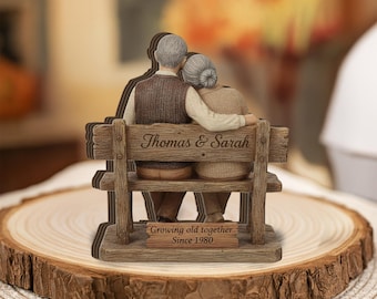 May include: A wooden figurine of an elderly couple sitting on a bench, with the names "Thomas & Sarah" engraved on the backrest. The base reads "Growing old together Since 1980". The figurine is set on a wooden slice.