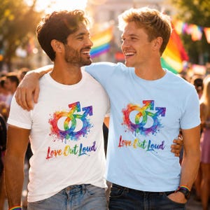 Puede incluir: Una camiseta blanca y una camiseta azul claro, cada una con un gráfico de doble símbolo de género masculino en estilo acuarela colorido y el texto "Love Out Loud" impreso en una fuente vibrante con degradado de arcoíris en el pecho.