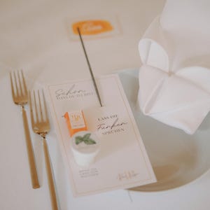 May include: A table setting with gold forks, a folded white napkin, and a card with text. The card has a small orange box and a white cup with a green leaf. A sparkler is placed on the card.