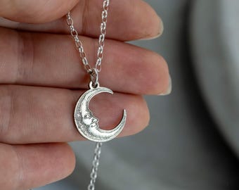 May include: A silver-colored necklace featuring a crescent moon pendant with a smiling face. The pendant is suspended from a delicate chain. The necklace is held up against a neutral background.