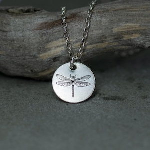 Può includere: Una collana color argento con un ciondolo rotondo con un design dettagliato di libellula. La libellula è incisa in nero, con ali e corpo delicati. La catena è anch'essa argentata. Il ciondolo ha un diametro di circa 2,5 cm.