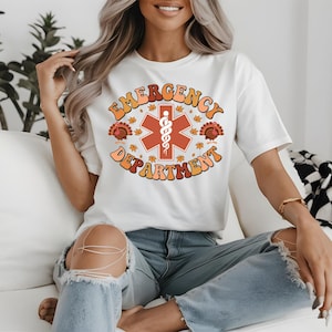 May include: White t-shirt with a retro design that says "Emergency Department" in orange and brown lettering. The design includes a red medical symbol, turkeys, and fall leaves. The shirt is worn by a person.