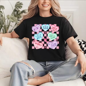 May include: Black t-shirt featuring a checkerboard design with pink and black squares. The shirt has heart-shaped graphics with phrases like "XOXO" and "UR SUCH A QT". The design includes pastel colors and text.