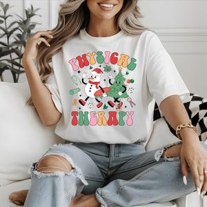 May include: White t-shirt with a colorful graphic that reads "PHYSICAL THERAPY". The design features a snowman and a Christmas tree, both with arms and legs, walking together. The shirt is a crew neck and has short sleeves.