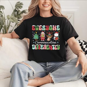 Op de afbeelding: Zwart T-shirt met de tekst "EVERYONE COMMUNICATION DIFFERENTLY" in kleurrijke letters. Het shirt toont kerst-thema cartoonfiguren, waaronder een kerstboom, de kerstman, een rendier en een sneeuwpop.