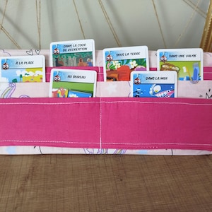 Könnte beinhalten: Ein rosa Stoff-Organizer mit mehreren Taschen, der illustrierte Karten mit französischem Text enthält. Der Organizer hat ein kontrastierendes rosa Band auf der Vorderseite und ist mit einem hellrosa Muster verziert.