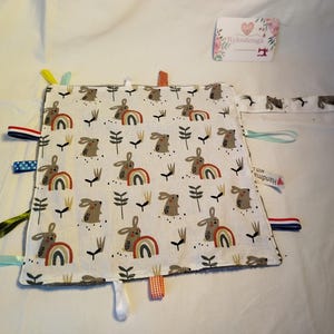 Puede incluir: Una manta de seguridad para bebé blanca con un estampado repetido de conejitos marrones, arcoíris y plantas verdes. La manta tiene cintas de colores y un cierre de botón a presión. Una etiqueta en la esquina dice "Handmade with love".