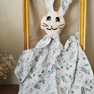 May include: A cream-coloured bunny comforter with long ears and a floral pattern. The bunny has black embroidered eyes and whiskers. The blanket has a white background with blue and green leaf designs.