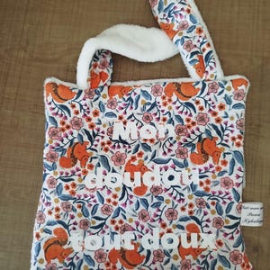 Puede incluir: Un bolso tote blanco con un estampado floral y de ardillas, con orejas y asas de conejo blancas de felpa. La bolsa tiene las palabras "Mon doudou tout doux" en letras blancas. Una pequeña etiqueta está adjunta al costado.