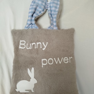 Puede incluir: Un bolso tote de felpa color topo con orejas de conejo y las palabras "Bunny power" en blanco. Las orejas de conejo son de cuadros azules claros y blancos. Una silueta de conejo blanco está en la parte inferior izquierda de la bolsa.