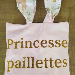 Puede incluir: Un bolso tote de felpa rosa con orejas de conejo. Las palabras "Princesse paillettes" están escritas en purpurina dorada. Las orejas de conejo son blancas con un estampado floral y un interior rosa.