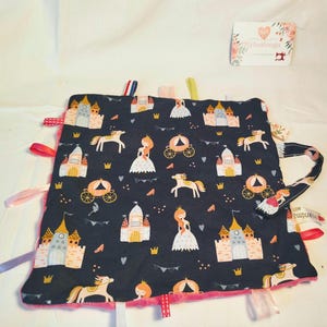 May include: A navy blue baby blanket with a princess theme. The blanket features illustrations of castles, carriages, princesses, and horses. It has a pink plush backing and colorful ribbons around the edges. A small tag with floral design is in the upper right corner.