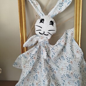 May include: A white bunny-shaped baby comforter with a floral pattern in blue and brown. The bunny has long ears with the same floral pattern. The comforter is hanging in a gold-coloured frame.