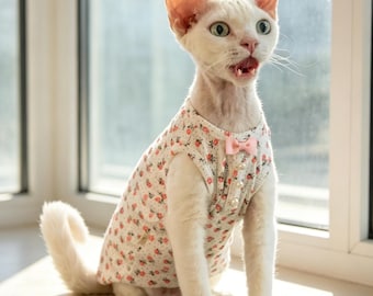 May include: A white cat with large ears and a short coat wearing a sleeveless shirt featuring a small pink floral pattern on a white background, accented with a tiny pink bow at the center of the neckline and a vertical row of small white buttons.
