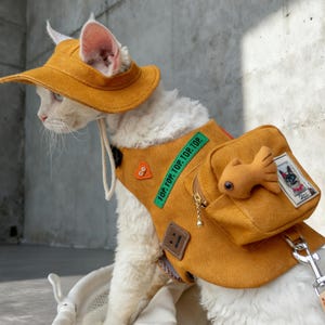 以下が含まれることがあります： マスタードイエローの帽子と、小さなバックパックが付いたお揃いのベストを着た白い猫。 ベストには「TOP. TOP. TOP. TOP.」の文字と、小さな魚の形をしたアクセサリーが付いています。 猫は白いバスケットの中に座っています。