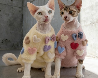 May include: Two Sphynx cats wearing matching sweaters. One cat is in a yellow sweater with heart and bow accents, the other in a pink sweater with similar details. The cats are sitting in front of a gray concrete wall.