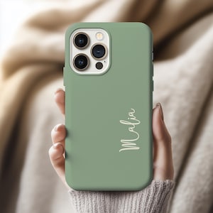 May include: Sage green phone case with the name "Malia" in a cursive font. The case has a smooth finish and a camera cutout. The phone case is held by a person.