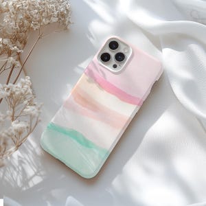 May include: A pastel-colored phone case with a watercolor design. The case features a soft pink, peach, and mint green color palette. The phone case is lying on a white surface with dried flowers in the background.