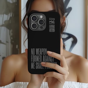 May include: Black phone case with the text "NO WEAPON FORMED AGAINST ME SHALL PROSPER" and "ISAIAH 54:17" in a light gray font. The case has a camera cutout and is held in front of a person.
