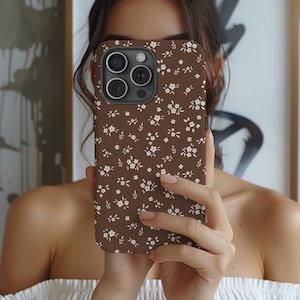 May include: A brown phone case with a white floral pattern. The case has a matte finish and features a repeating design of small white flowers and leaves. The phone case is designed to fit a smartphone.