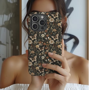 May include: A phone case with a dark background featuring a floral and butterfly pattern in shades of beige, brown, and white. The case has a camera cutout and is held up in front of a person.