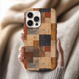 May include: A phone case with a patchwork design in shades of brown, tan, and black. The case has a textured appearance and is held in a person's hand. The phone's camera lenses are visible.