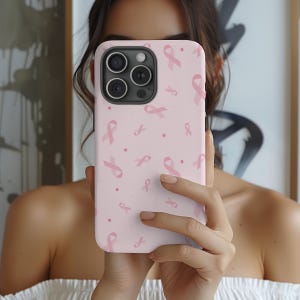 Könnte beinhalten: Eine hellrosa Handyhülle mit einem Muster aus rosa Awareness-Schleifen und kleinen Punkten. Die Hülle wird vor einer Person gehalten, die ihr Gesicht verdeckt. Die Handyhülle hat ein Kameramodul mit drei Linsen.