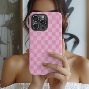 May include: A pink and white checkered phone case. The case is held up in front of a person's face, obscuring the face. The phone case has a black camera module.