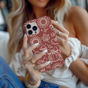 May include: A maroon phone case featuring a fall-themed design. The case is adorned with white illustrations of sunflowers, leaves, pumpkins, mushrooms, and acorns. The phone case is held by a person with long blonde hair and wearing rings.