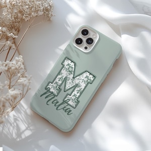 May include: A sage green phone case featuring the letter "M" in a floral design. The name "Malia" is written in a script font below the letter. The case has a smooth finish and is designed for a smartphone.