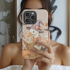 May include: A beige phone case featuring a whimsical Halloween illustration. It depicts a goose wearing a witch hat and scarf, reading a book, with pumpkins, ghosts, and a black cat. The case has a camera cutout and a smooth finish.